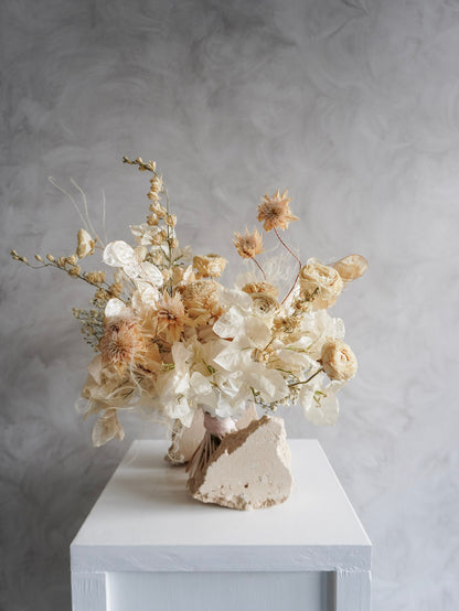 A dried flower bridal bouquet positioned against a rough, natural stone, featuring cream and soft beige florals, displayed on a white plinth against a textured gray background.