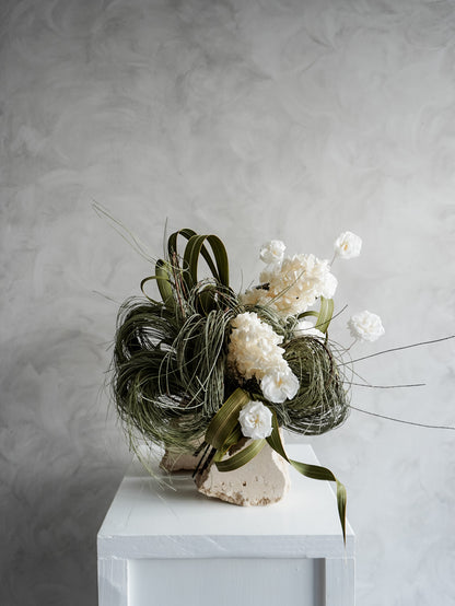 Bridal bouquet of dried white flowers and greenery on a white plinth against a gray background.