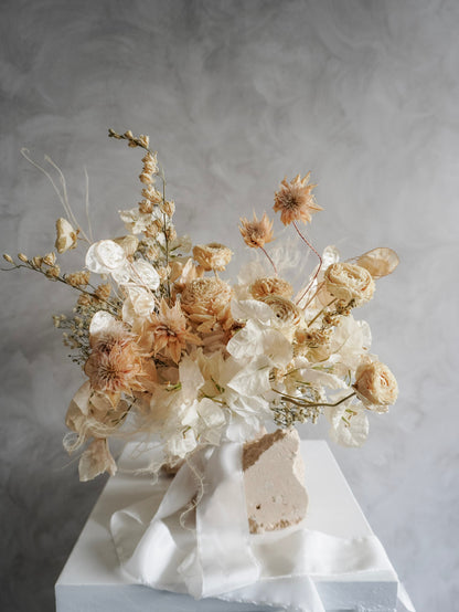 A dried flower bridal bouquet positioned against a rough, natural stone, featuring cream and soft beige florals with draping white ribbon, displayed on a white plinth against a textured gray background.