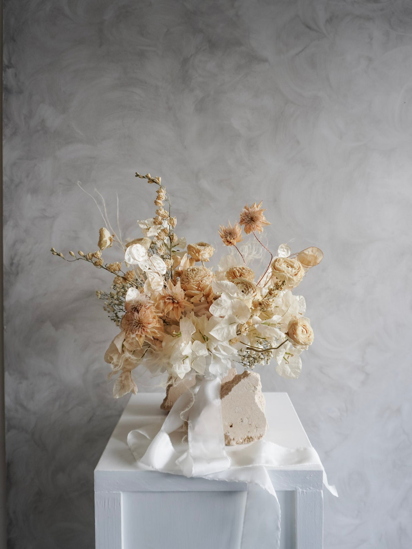 A dried flower bridal bouquet positioned against a rough, natural stone, featuring cream and soft beige florals with draping white ribbon, displayed on a white plinth against a textured gray background.