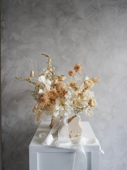 A dried flower bridal bouquet positioned against a rough, natural stone, featuring cream and soft beige florals with draping white ribbon, displayed on a white plinth against a textured gray background.