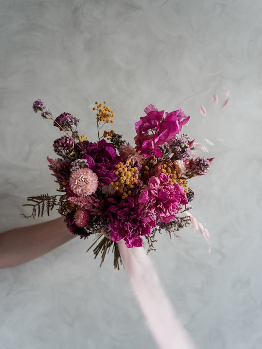 Bridal bouquet of dried pink and purple flowers held against a neutral background.