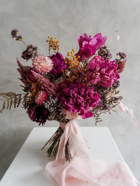 Bridal bouquet of dried pink and purple flowers on a white plinth with a gray background.