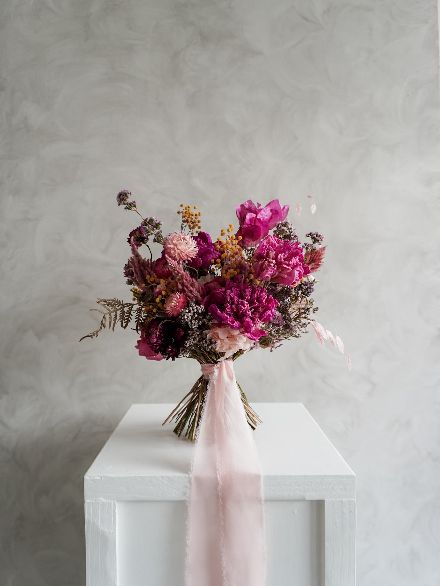 Bridal bouquet of pink and purple flowers on a white plinth against a gray background.