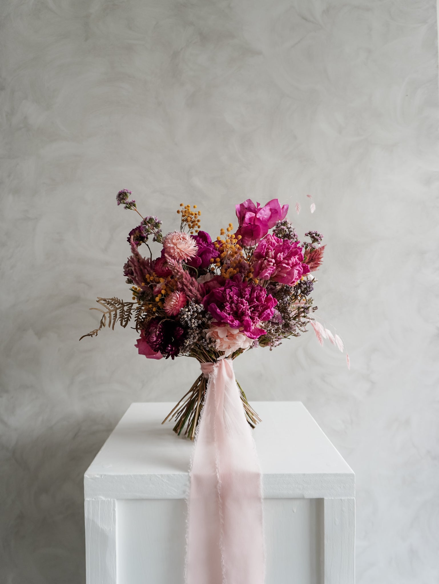 Bridal bouquet of pink and purple flowers on a white plinth against a gray background.