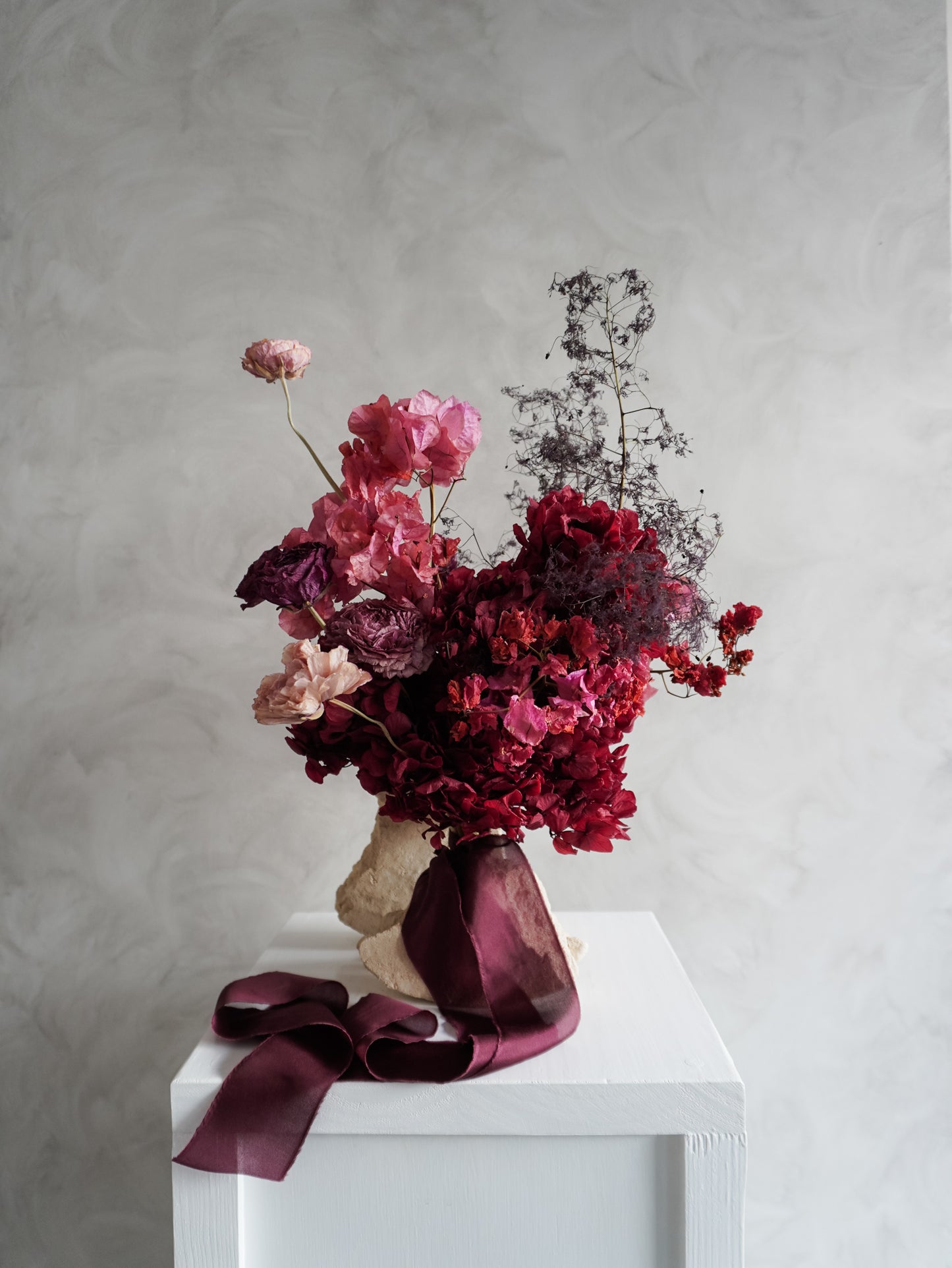 Bridal bouquet of dried and preserved pink and burgundy flowers on a white plinth in front of a gray background.