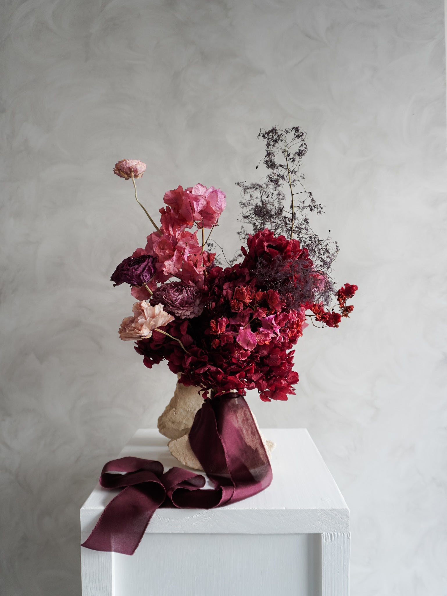 Bridal bouquet of dried and preserved pink and burgundy flowers on a white plinth in front of a gray background.