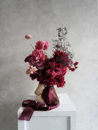 Bridal bouquet of dried and preserved pink and burgundy flowers on a white plinth in front of a gray background.