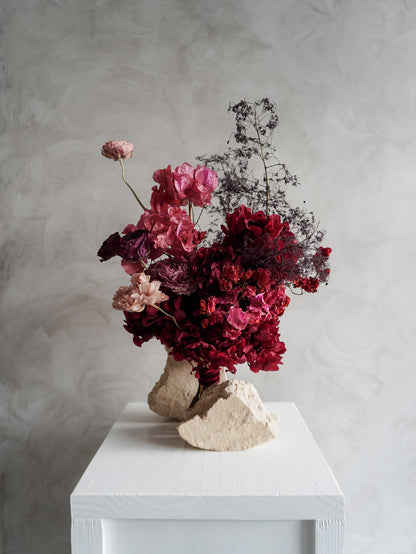 Bridal bouquet of dried and preserved pink and burgundy flowers on a white plinth in front of a gray background.