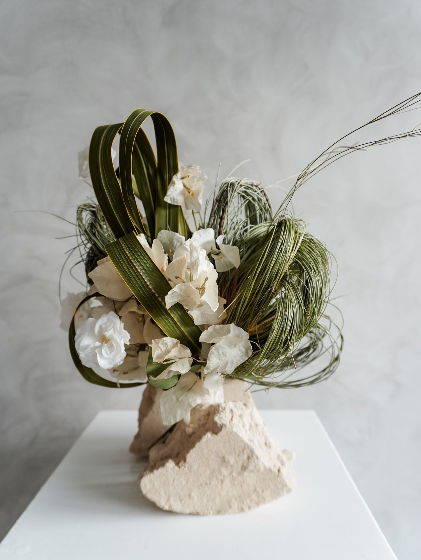 Bridesmaid bouquet of preserved white roses and greenery on a white plinth against a plain background.