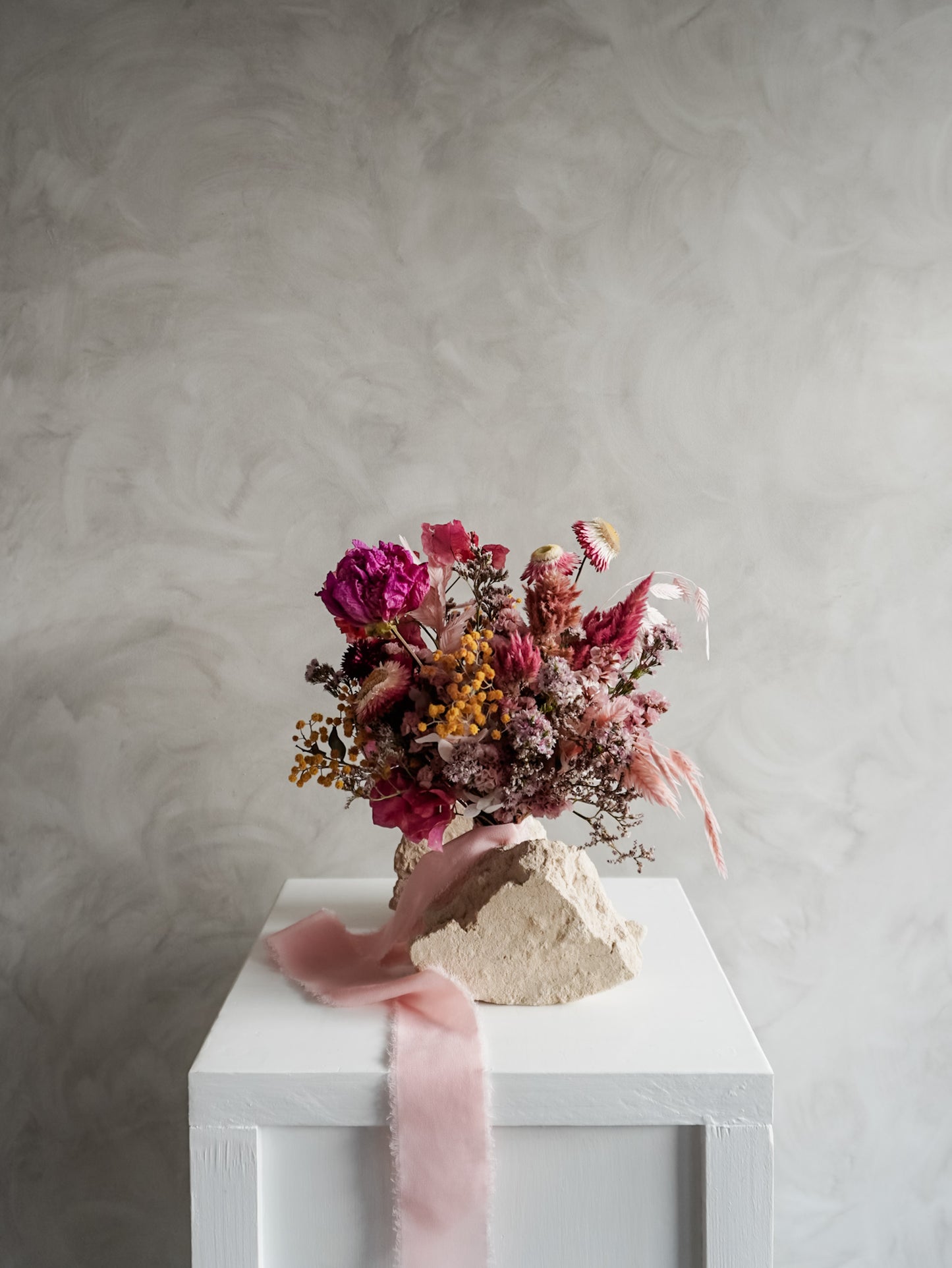 Bridesmaid bouquet of dried flowers on a white plinth with a pink ribbon against a gray textured wall.