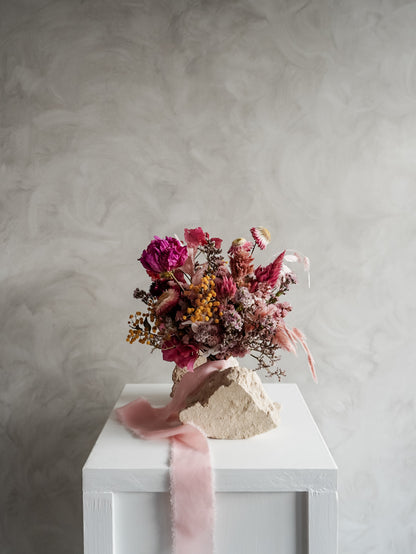 Bridesmaid bouquet of dried flowers on a white plinth with a pink ribbon against a gray textured wall.