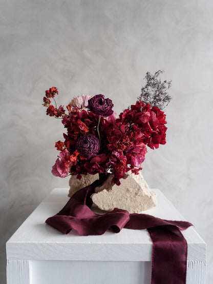 Bridesmaid bouquet of dried red and purple flowers on a white plinth with a gray background.