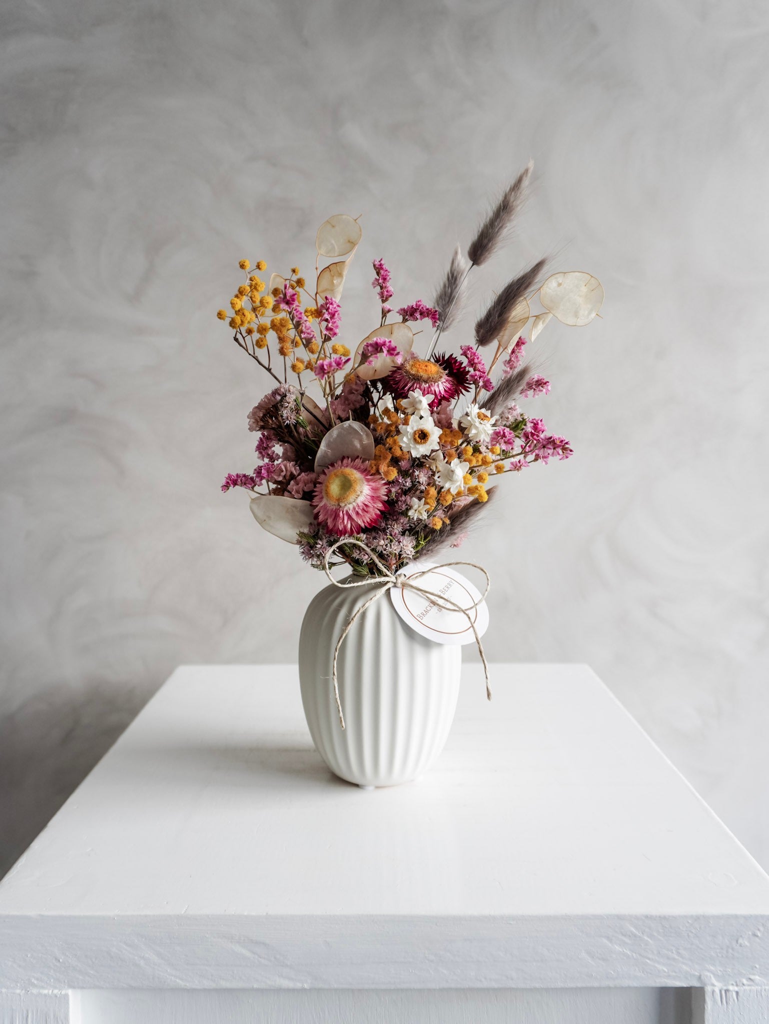 Dried flower arrangement in bright colours displayed in a white bud vase and positioned on a white plinth against a gray background. Distant view.