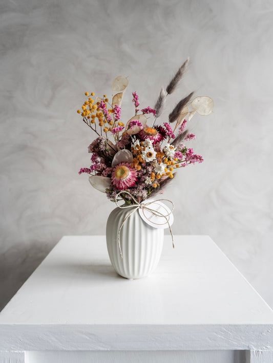Dried flower arrangement in bright colours displayed in a white bud vase and positioned on a white plinth against a gray background. Distant view.