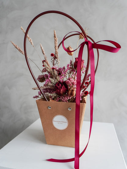Dried floral arrangement in a kraft posy bag with a red ribbon on a white surface.