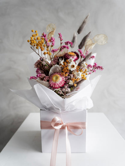 Dried flower arrangement in bright colours displayed in a white posy box and positioned on a white plinth against a gray background.