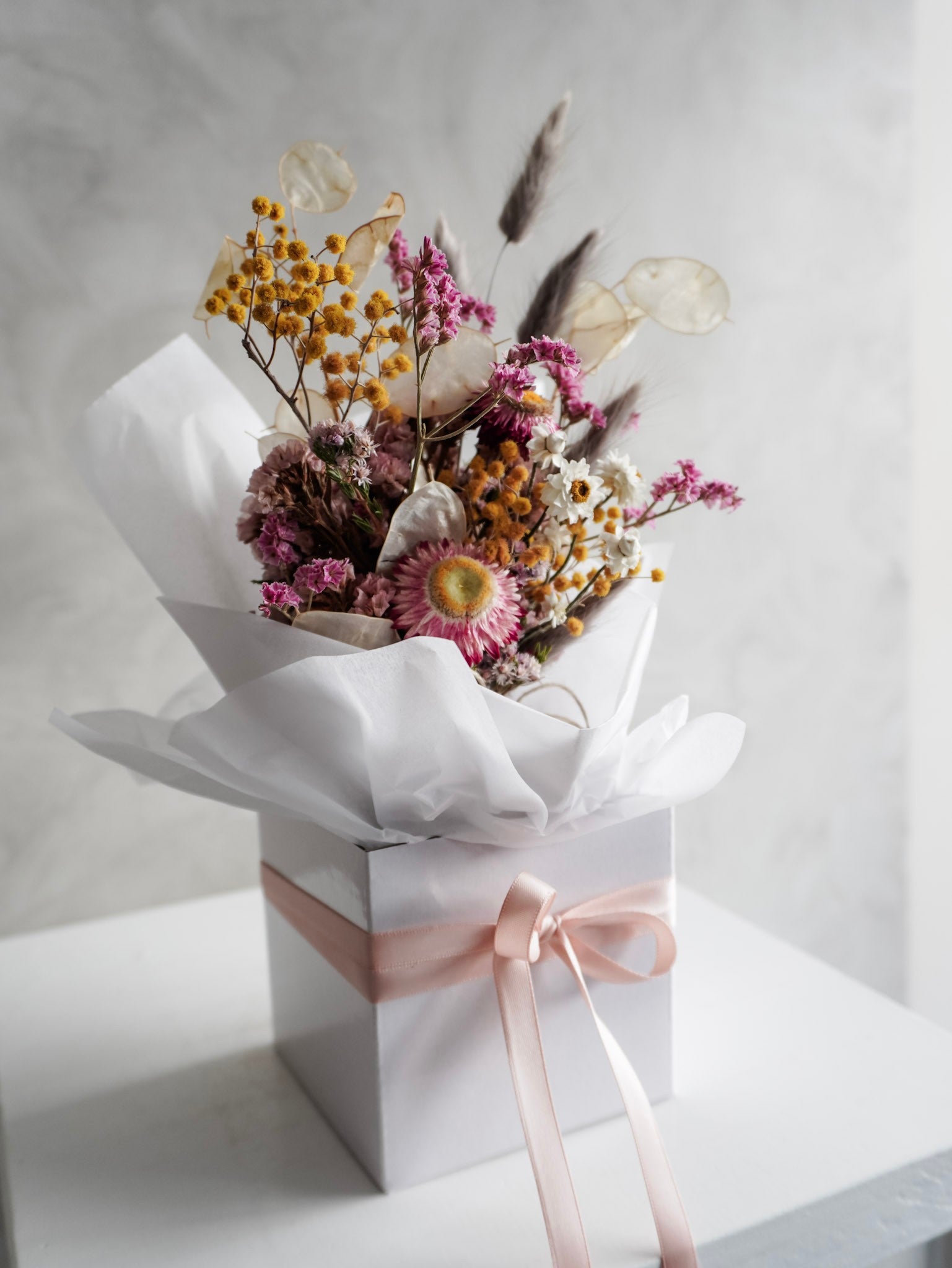 Dried flower arrangement in bright colours displayed in a white bud vase and positioned on a white plinth against a gray background. Side view.
