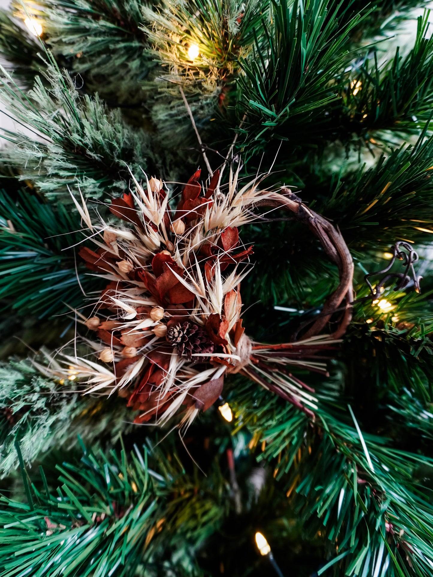 Dried flower mini wreath decoration in earthy tones on a grapevine base hung on a green Christmas tree.