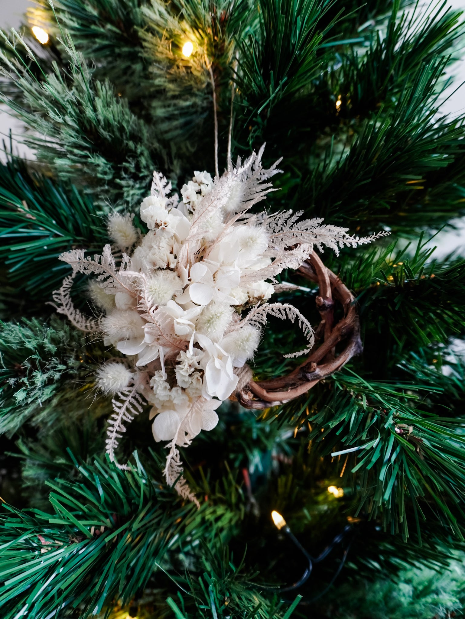 Dried flower mini wreath decoration in soft pink and white tones on a grapevine base hung on a green Christmas tree.