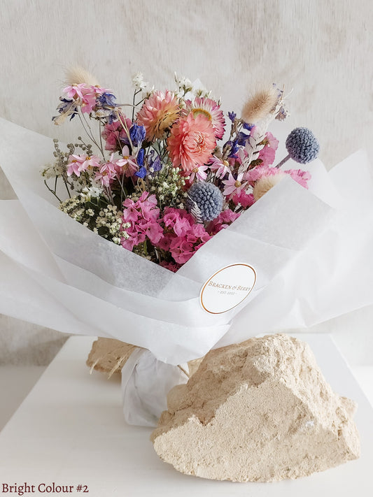 A petite dried flower bouquet wrapped in white tissue paper, featuring bright pink, blue, and white flowers on a white plinth against a light gray background.