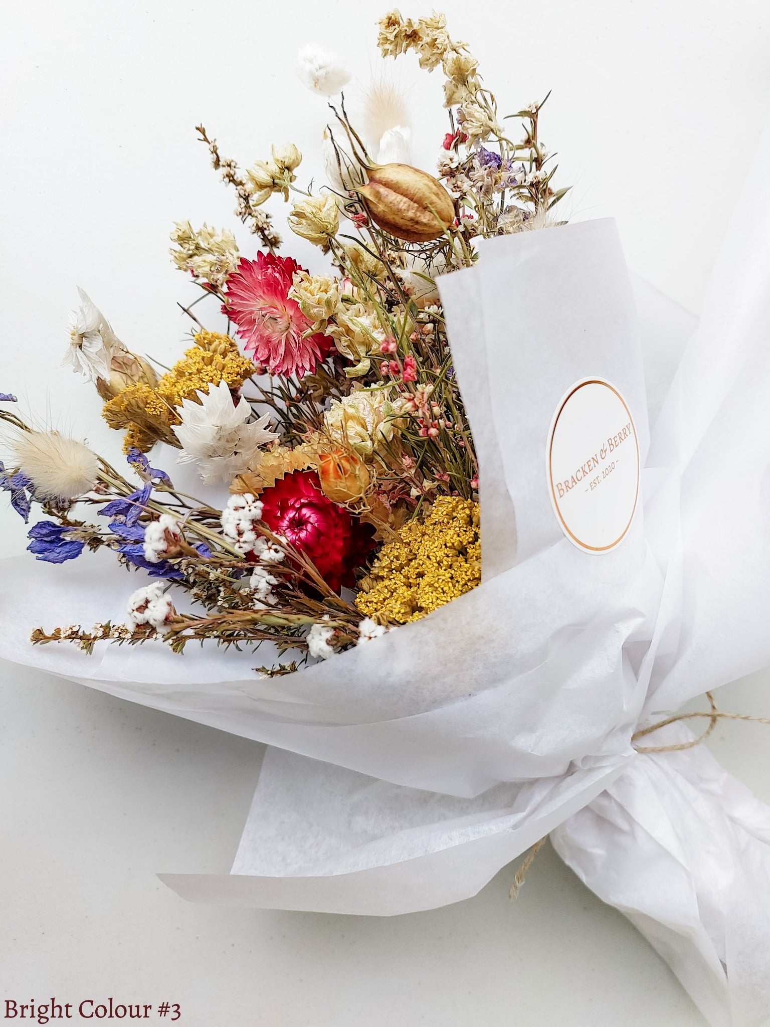 A petite dried flower bouquet wrapped in white tissue paper, featuring pink, and yellow flowers on a white plinth against a white table.
