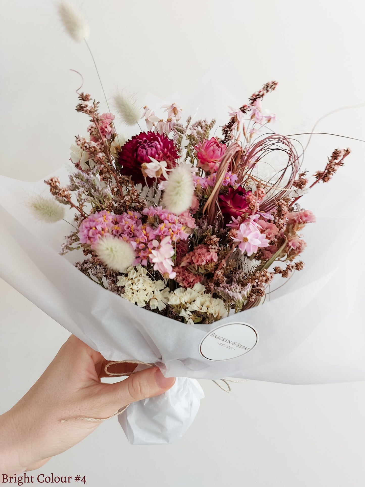 A dried flower bouquet in white tissue paper, held by a hand against a plain white background. The bouquet features pink, burgundy, and cream dried flowers and grasses.