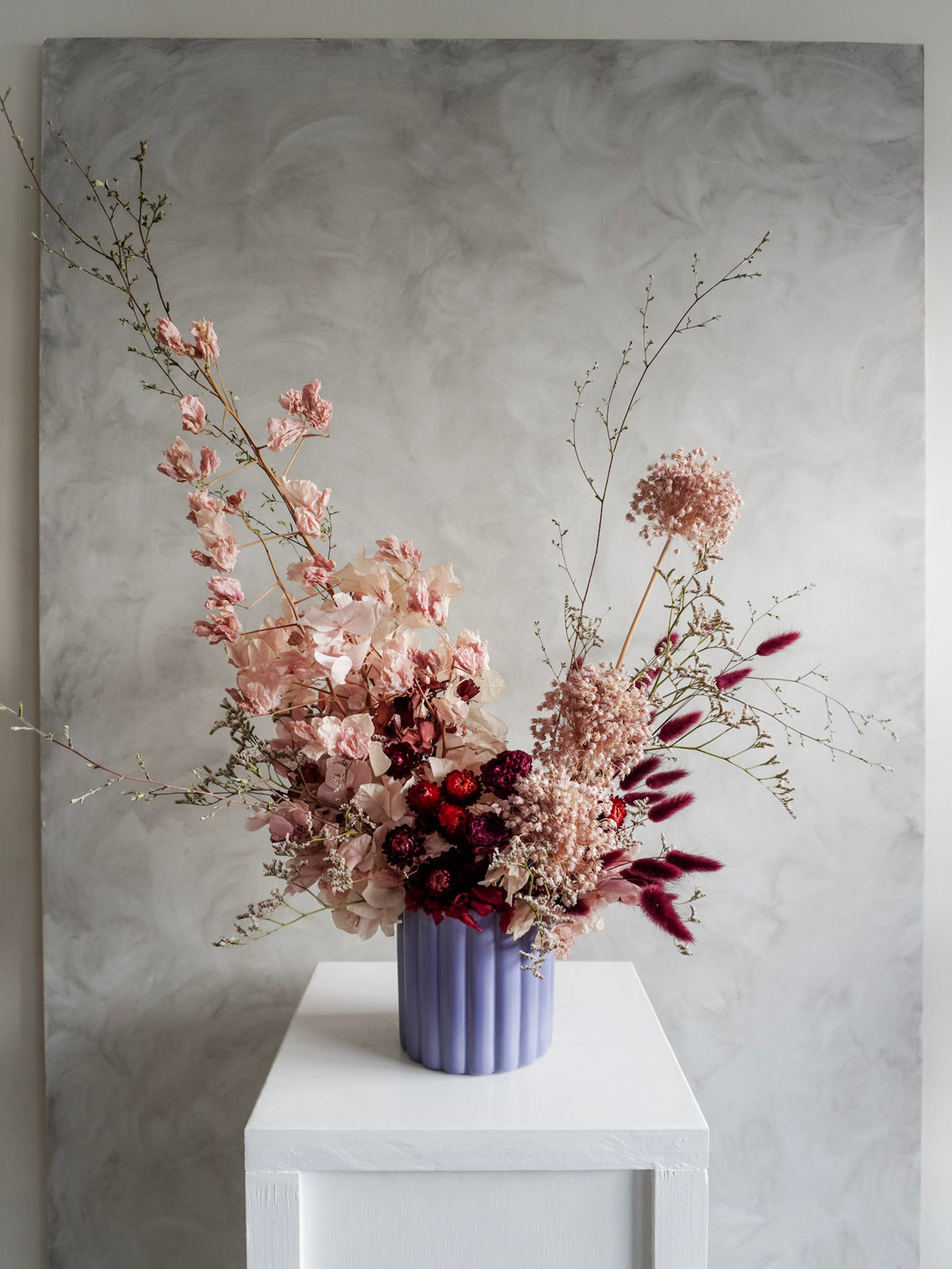 A dried flower arrangement in a lavender ribbed ceramic vase, displayed on a white plinth against a textured gray background.