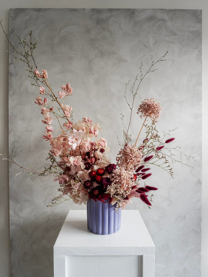 A dried flower arrangement in a lavender ribbed ceramic vase, displayed on a white plinth against a textured gray background.