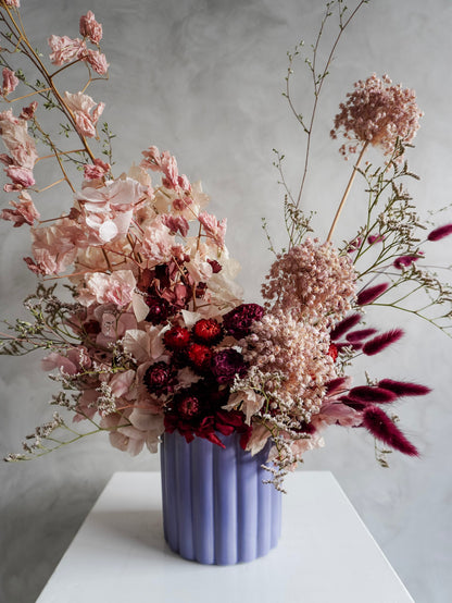 A dried flower arrangement in a lavender ribbed ceramic vase, featuring dusty pink, burgundy, and deep purple botanicals on a white plinth against a textured gray background.