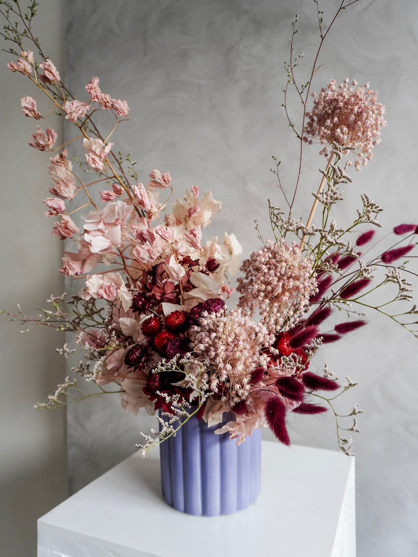 A dried floral arrangement in a lavender ribbed ceramic vase, featuring dusty pink, burgundy, and cream botanicals, displayed on a white plinth against a textured gray background.