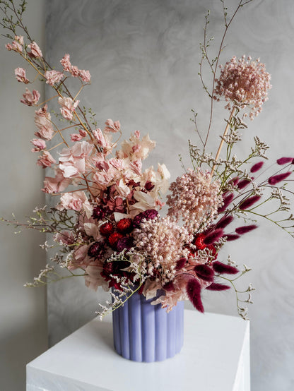 A dried floral arrangement in a lavender ribbed ceramic vase, featuring dusty pink, burgundy, and cream botanicals, displayed on a white plinth against a textured gray background.