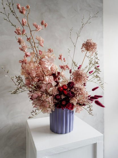 A dried flower arrangement in a lavender ribbed ceramic vase, displayed on a white plinth against a textured gray background.