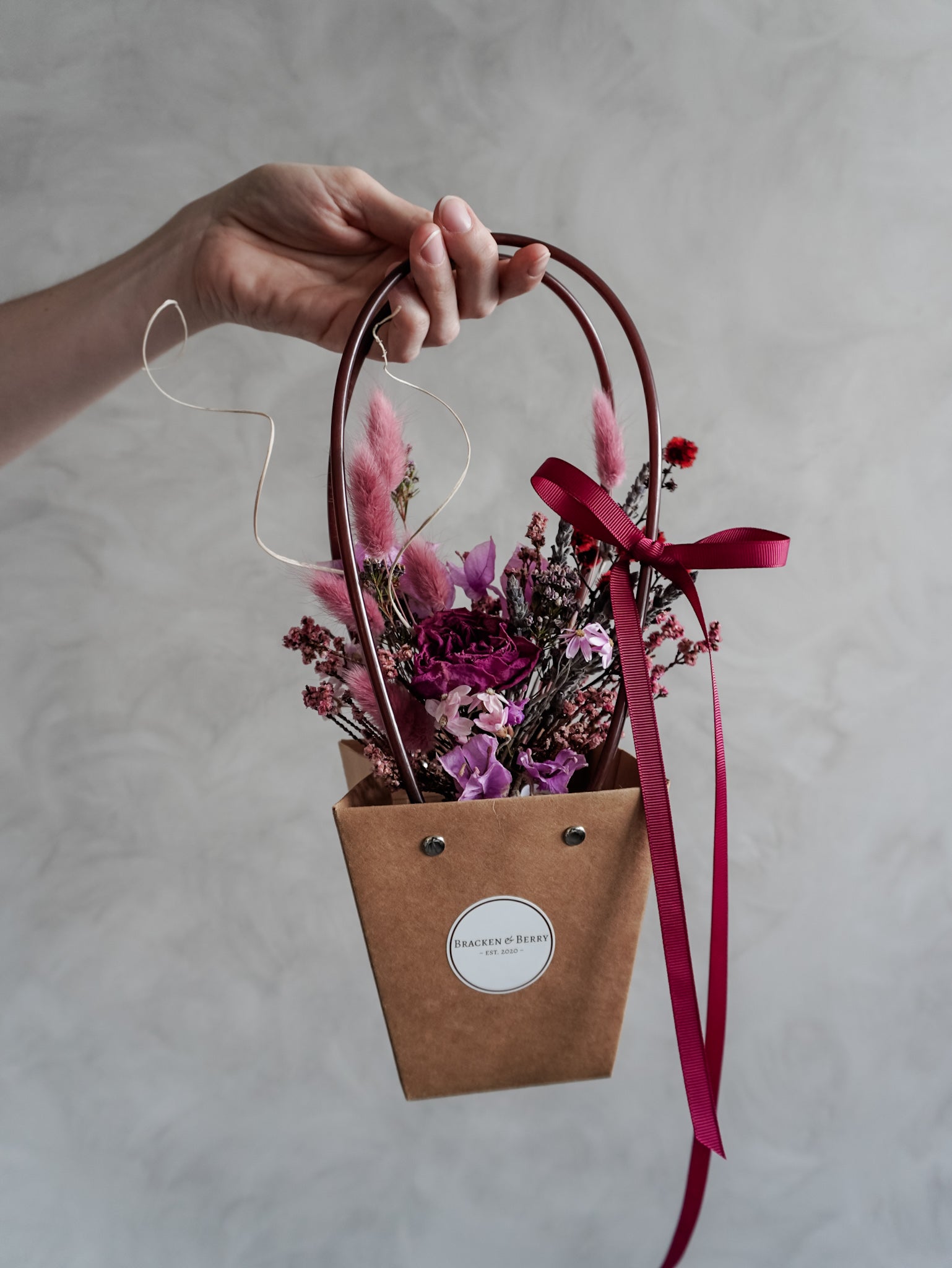 Hand holding a small brown posy bag with dried flowers and a pink ribbon against a neutral background