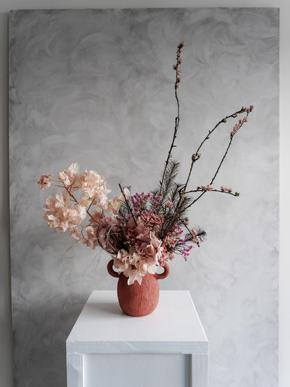 Urn vase with pink dried flowers on a white table against a gray wall.