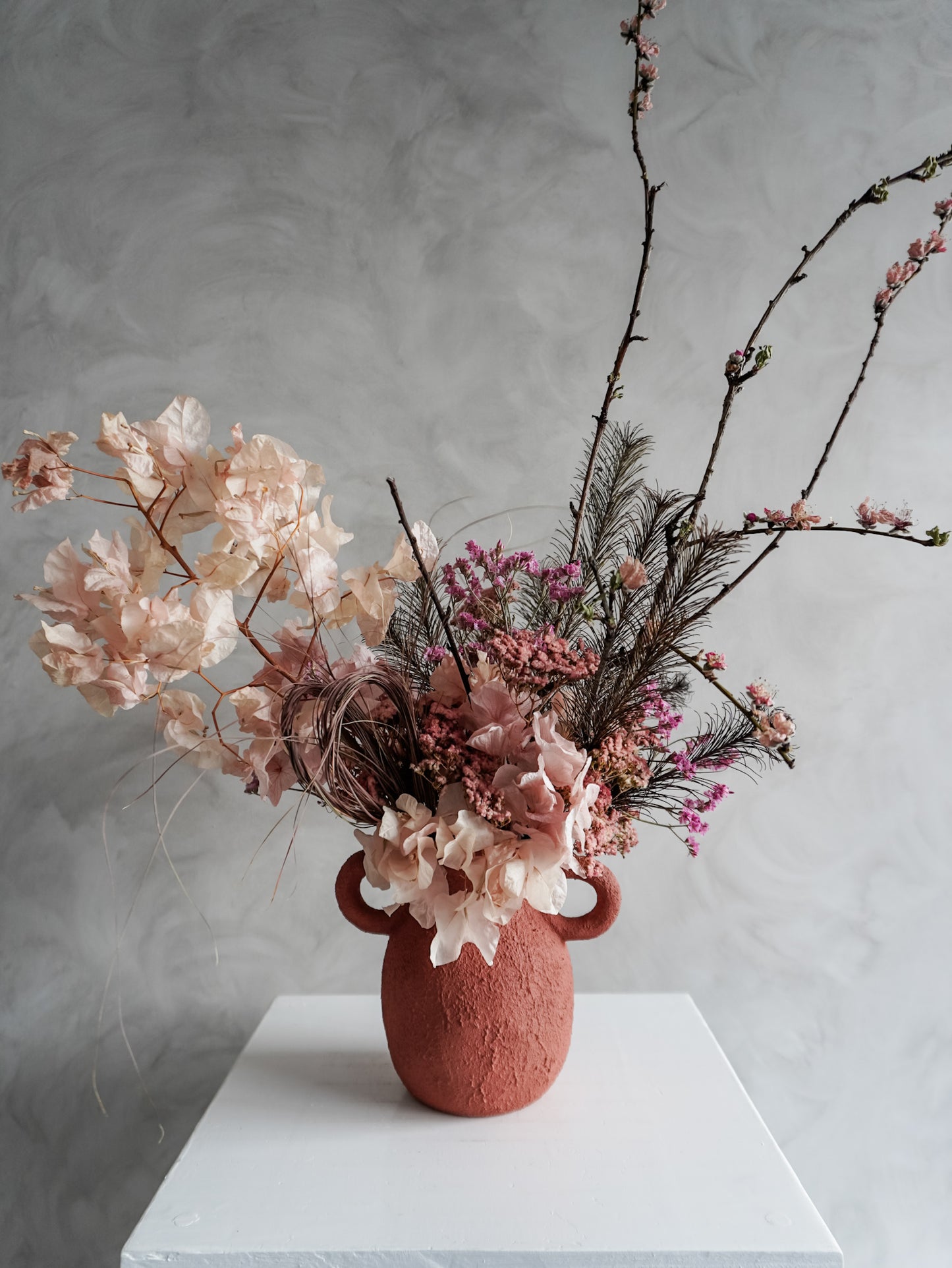 Textured urn vase with pink and burgundy dried flowers and branches on a white surface against a gray background.