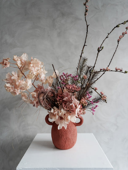 Textured urn vase with pink and burgundy dried flowers and branches on a white surface against a gray background.
