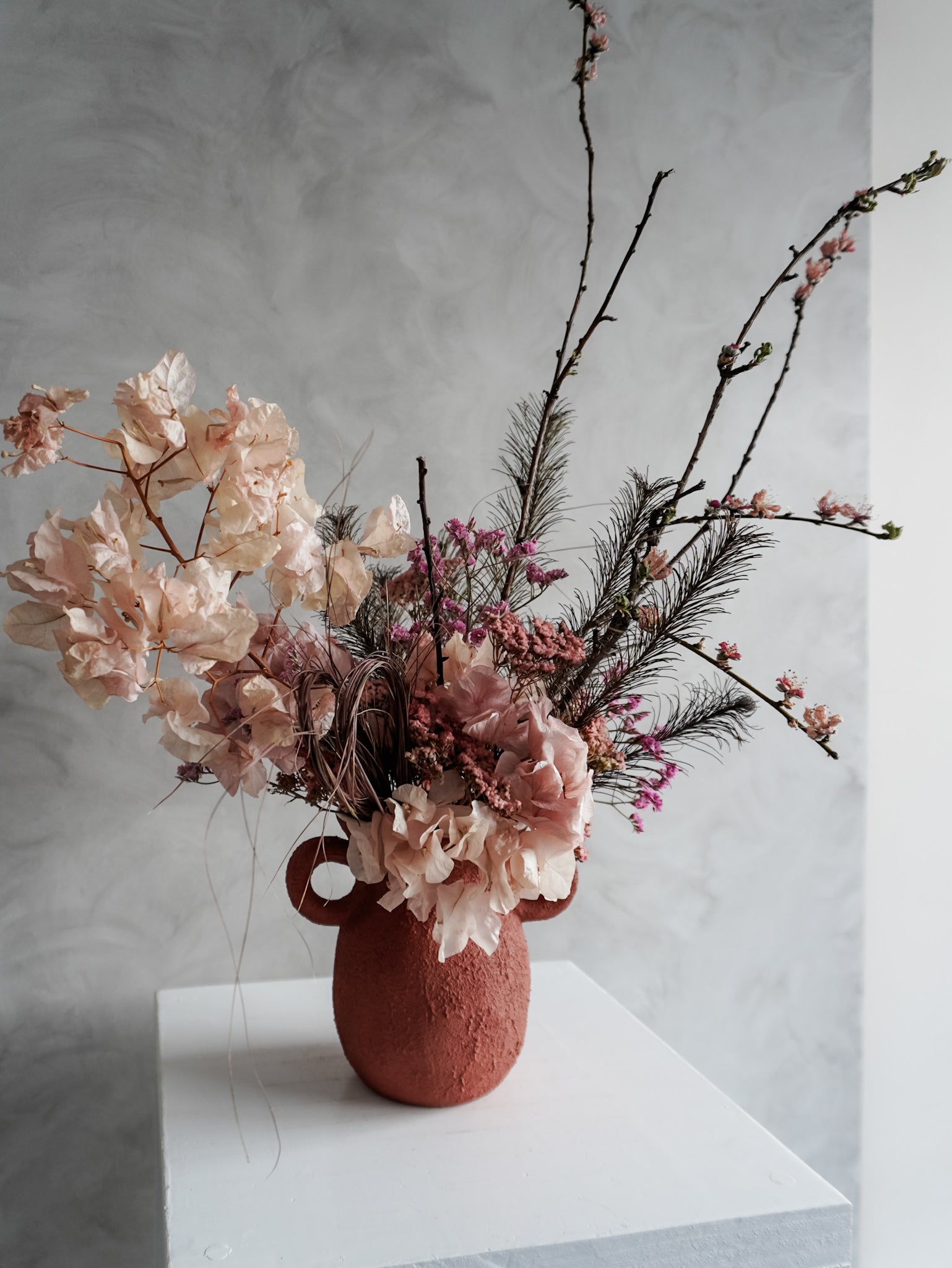 Pink dried floral arrangement in a terracotta urn vase on a white surface with a gray background.
