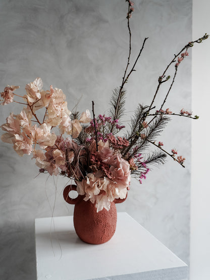 Pink dried floral arrangement in a terracotta urn vase on a white surface with a gray background.
