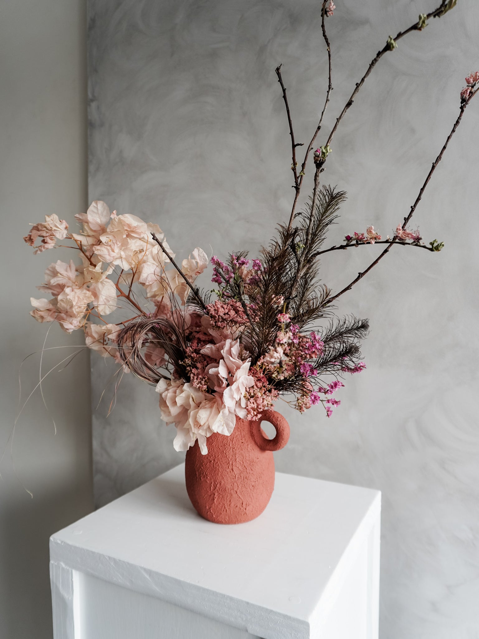 Pink dried floral arrangement in a terracotta urn vase on a white surface with a gray background.