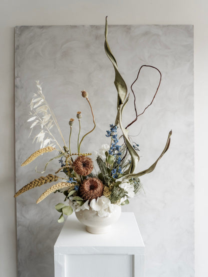 Dried flower arrangement in natural green and white tones in a white ceramic vase displayed on a white plinth against a gray background.