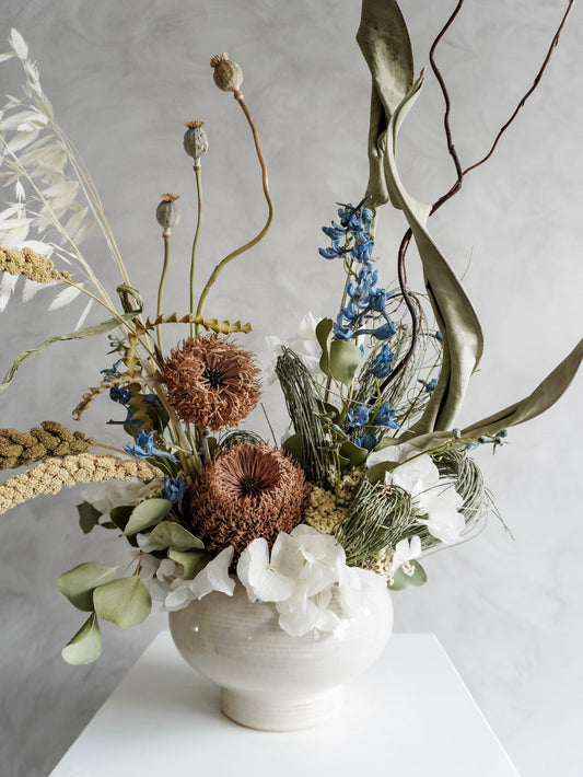 Dried flower arrangement in natural green and white tones in a white ceramic vase displayed on a white plinth against a gray background - close view.