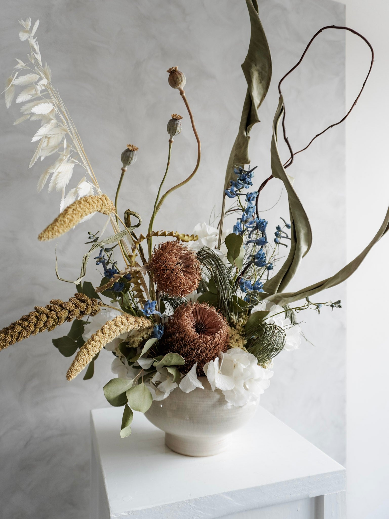 Dried flower arrangement in natural green and white tones in a white ceramic vase displayed on a white plinth against a gray background - side view.