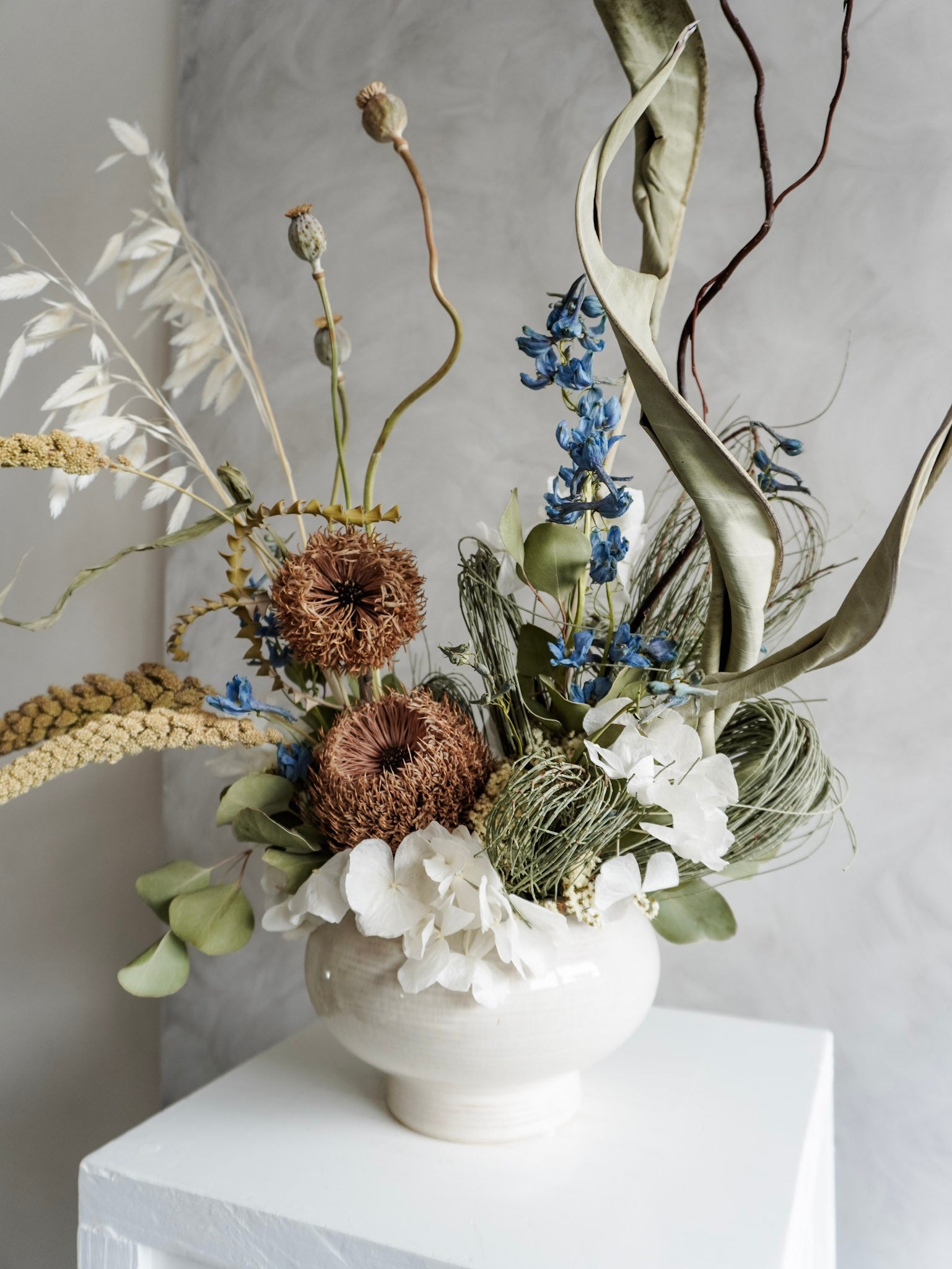 Dried flower arrangement in natural green and white tones in a white ceramic vase displayed on a white plinth against a gray background - side view.