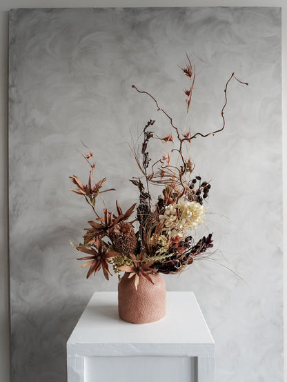 Dried floral arrangement in a textured vase on a white pedestal against a gray wall - distant view.