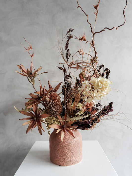 Dried floral arrangement in a textured vase on a white pedestal against a gray wall - close view.