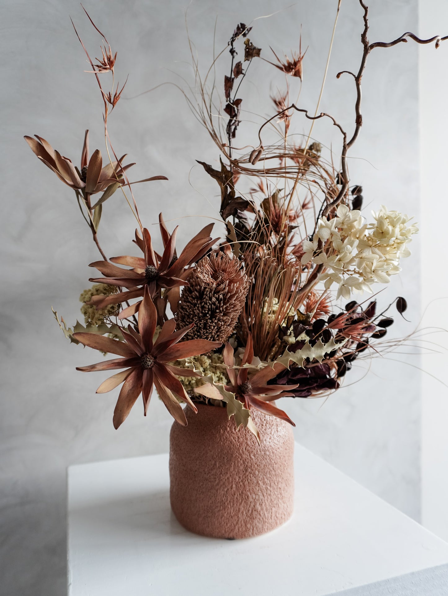 Dried floral arrangement in a textured vase on a white pedestal against a gray wall - side view.