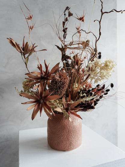 Dried floral arrangement in a textured vase on a white pedestal against a gray wall - side view.