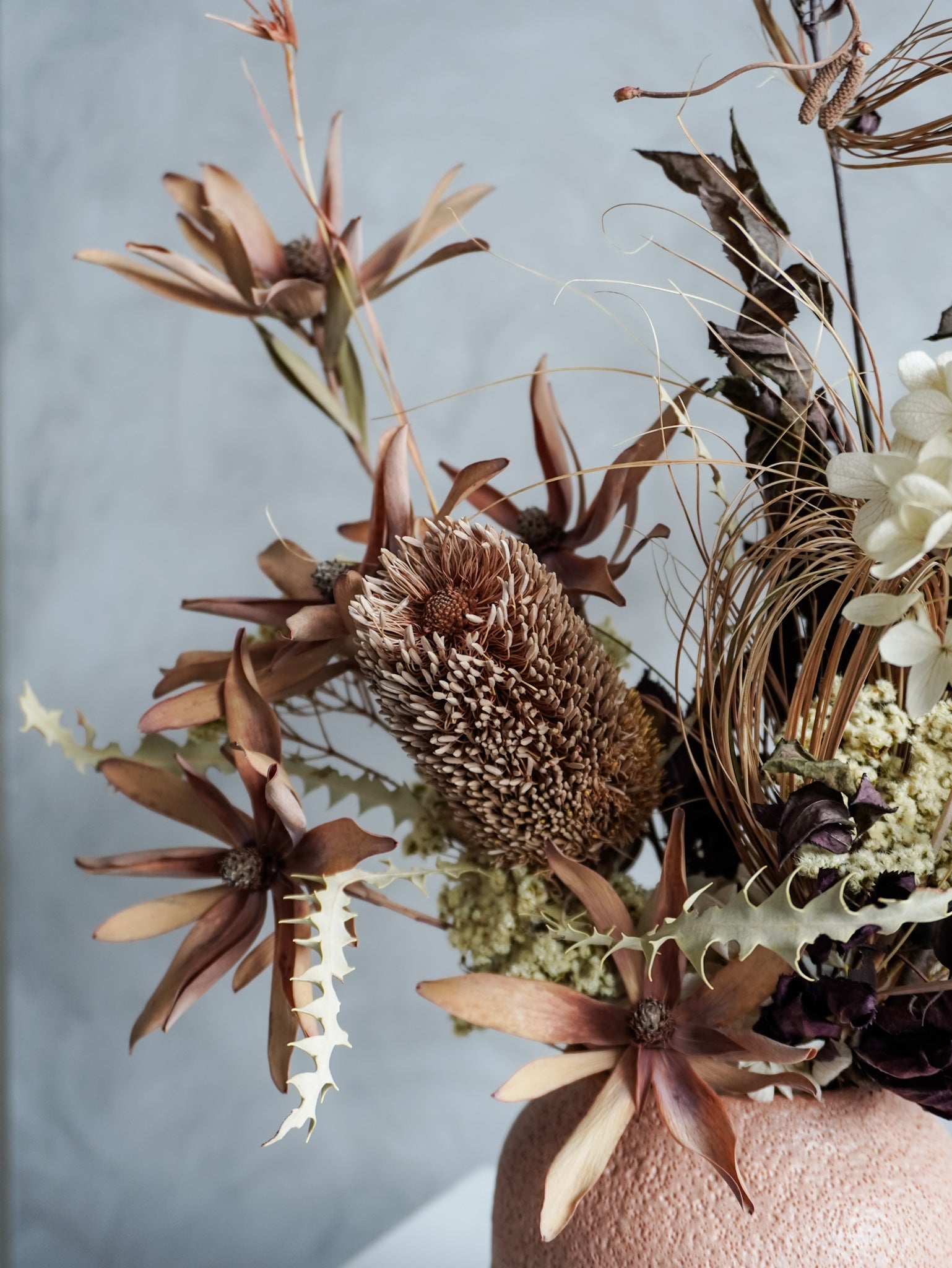 Close up of a dried floral arrangement in a textured vase against a gray wall.