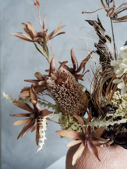 Close up of a dried floral arrangement in a textured vase against a gray wall.